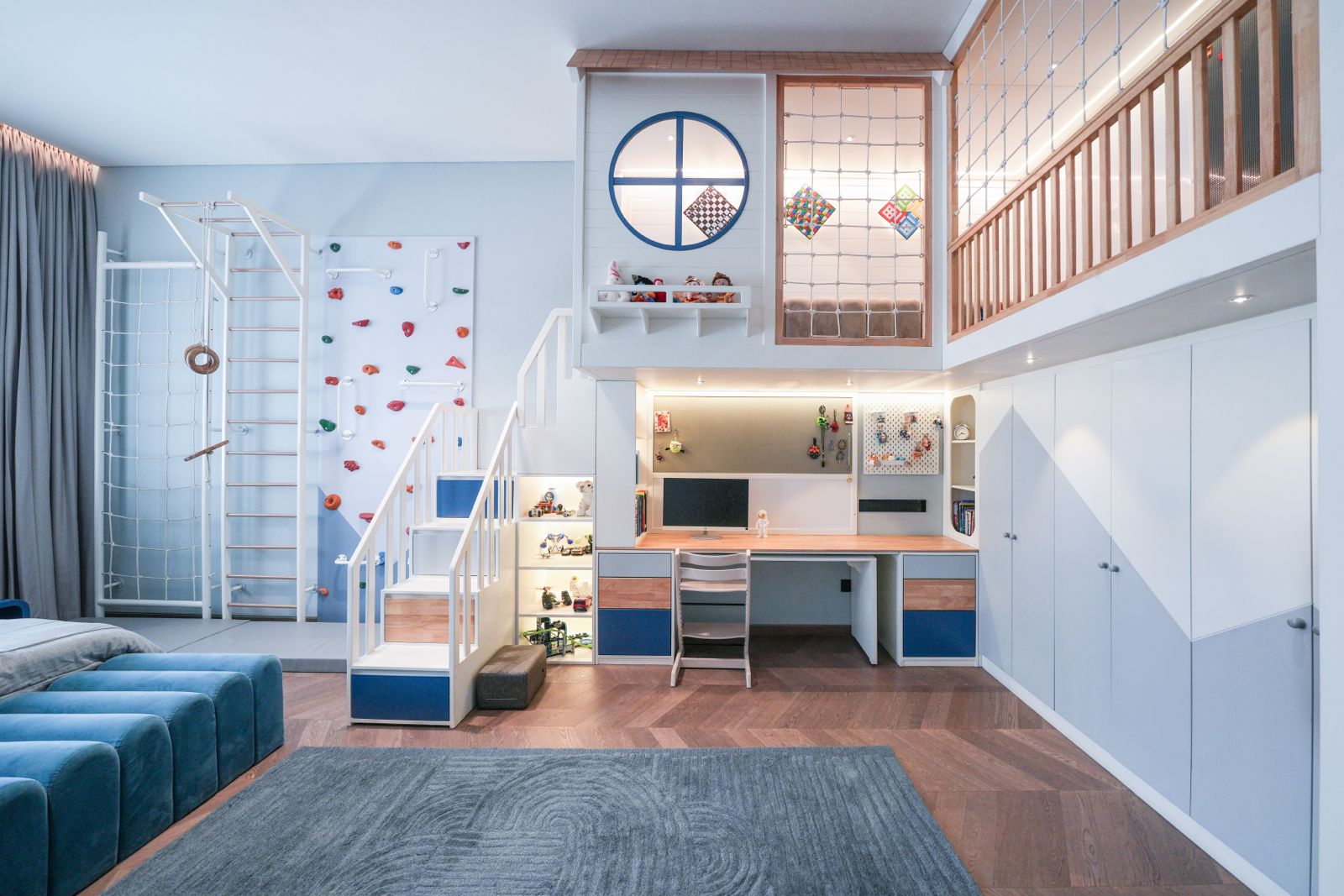 A completed Magical Nest children's room: a loft with a round window and rope netting above a built-in study desk, a climbing wall and wall bars on the left, and full-height wardrobes on the right.