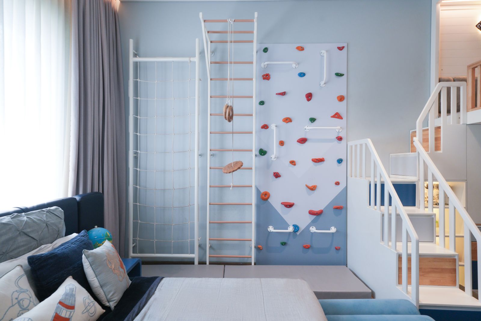 A pale blue wall fitted with a rope climbing net, wall bars with gymnastic rings and a swing seat, and a climbing panel with coloured holds above a padded mat, beside a white staircase to the loft.