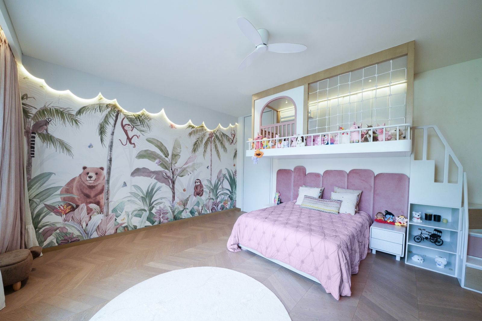 A completed Magical Nest room: a hand-painted jungle mural lit by a scalloped cove, a white play loft with rope netting and a staircase with shelves, above a bed with a tall upholstered pink headboard.