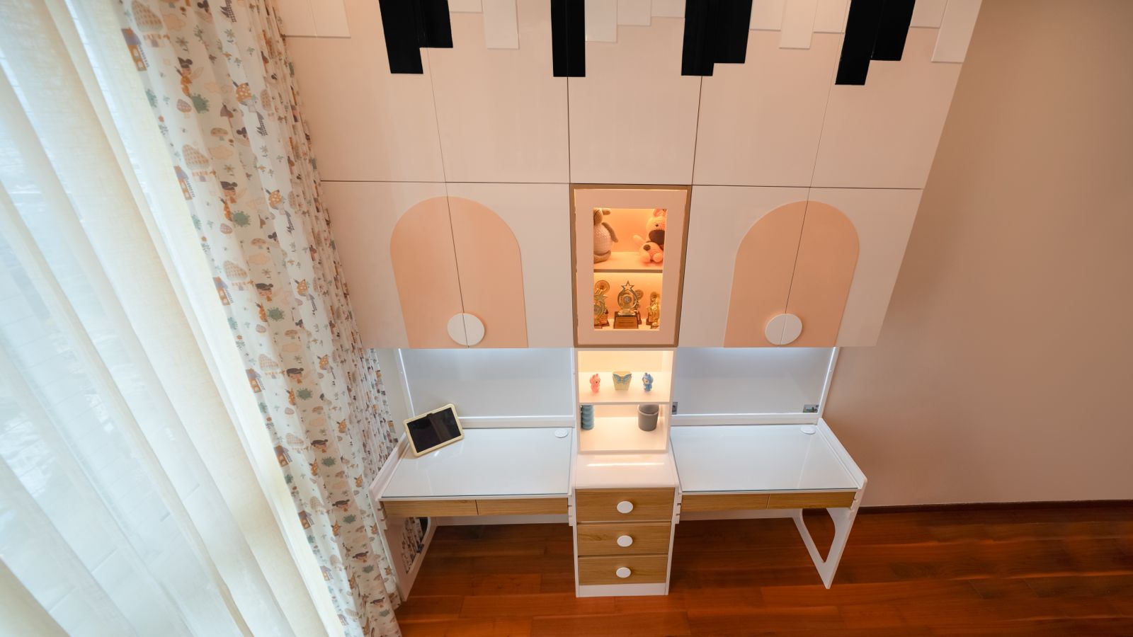 Twin white study desks with oak drawers either side of a lit display niche, beneath a wardrobe with piano-key panels and arched peach doors.